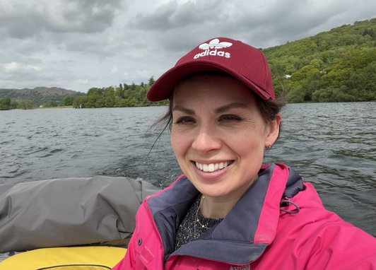 Trustee Natalie smiles from a small boat on what looks like a Loch. She is wearing a pink waterproof jacket and a burgundy cap.