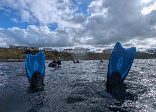 A low-angle, first-person view from the water of two blue swimming fins. In the background, several people are diving. A row of buildings under a partly cloudy sky can be seen behind them. The water is choppy and reflects the sunlight.