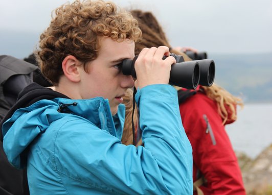 A young person looking through binoculars outside. They are wearing a light blue coat and there is someone standing behind htem wearing a red coat. In the background is the ocean.