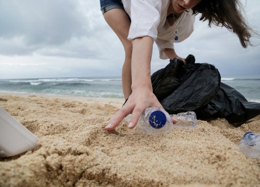 A female in a white top is reaching down to pick up a plastic bottle from the sand