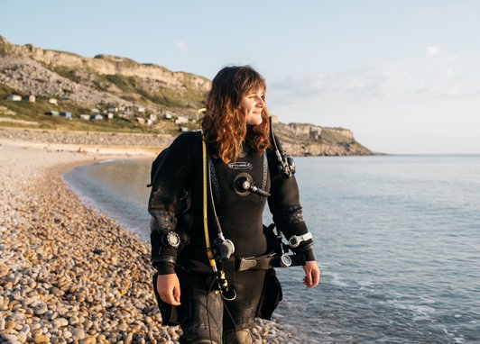 A woman wearing full black scuba diving gear, including a drysuit and a regulator, standing on a pebble beach at the water's edge, smiling and looking out towards the sea and a large cliff face in the background.