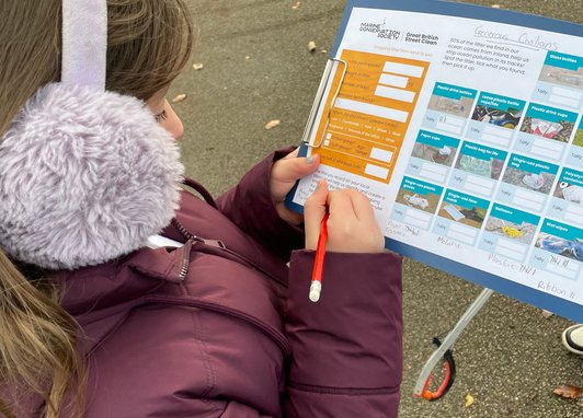A child wearing earmuffs and a purple coat is taking part in a Source to Sea survey. She is holding a form and writing on it with a red pencil.