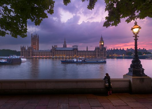 Westminster at sunset taken from the bank across the Thames. The sky is a stunning purple pink and an ornate lamp is lit in the foreground.