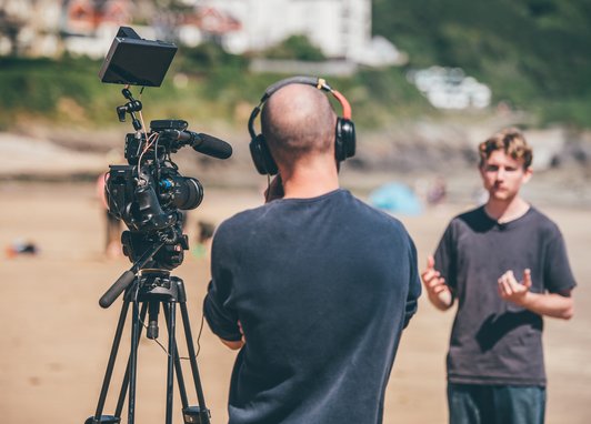 A videographer wearing headphones is operating a professional camera on a tripod on a sandy beach, while a young man stands nearby talking. The young man is blurred.
