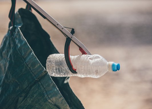 A close-up of a person using a litter picker tool to place a clear plastic water bottle with a blue cap into a dark green reusable bag. The person is out of shot.