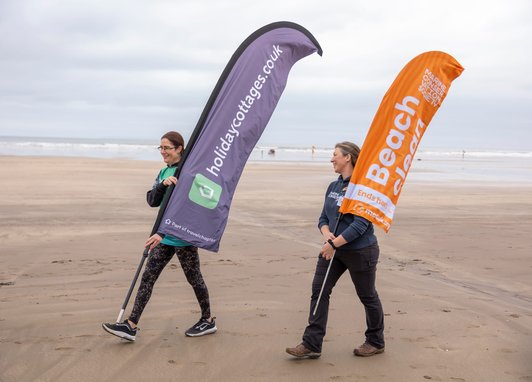 2 women walking along the beach carrying large marker flags. The flags read "Holidaycottages.co.uk" and "Marine Conservation Society Beach Clean" respectively