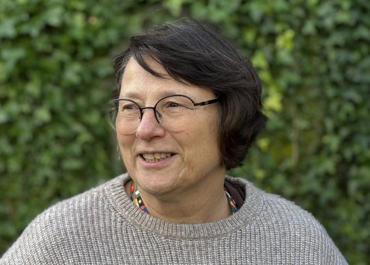 Mel stands sideways against a backdrop of a vivid green hedge. She wears a light grey jumper.