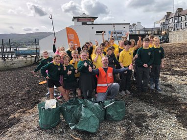 A group of primary school students standing on a beach. In the centre is an MCS employee wearing a hi-vis jacket. 5 dark green waste bags lay in front of them filled with litter found on the beach.