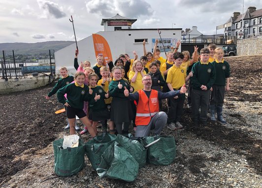 A group of primary school students standing on a beach. In the centre is an MCS employee wearing a hi-vis jacket. 5 dark green waste bags lay in front of them filled with litter found on the beach.