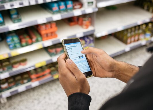 A person stands in front of the fish section in a supermarket holding a phone in their hand. On the phone screen is the Good Fish Guide species ratings page.
