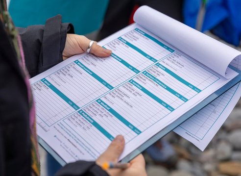 A close-up shot of a person holding a clipboard with a Marine Conservation Society beach clean survey form. The user's hands and a pen are visible as they potentially make notes on the form.