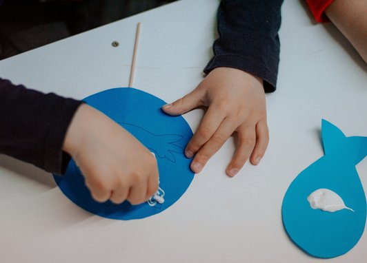the hands of a child making a fish out of blue paper, glue and a stick