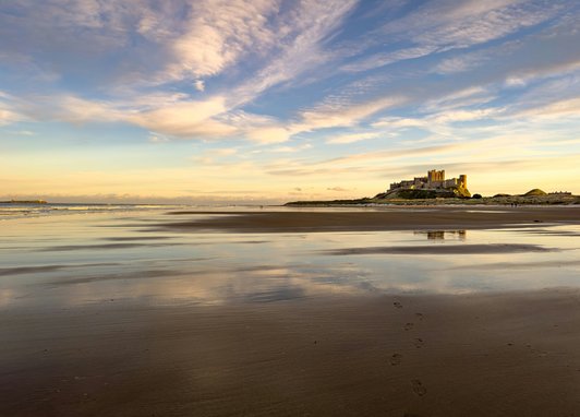 Bamburgh Castle as seen across Bamburgh Castle Beach. It looks like dusk, with a pale golden sky on the horizon and the colours reflecting off the water on the sand.