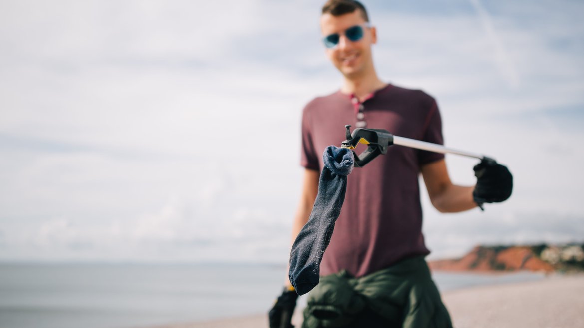 A man holding up a dark coloured sock up to the camera. He is using a litter picker to hold it. The sock is dark in colour and covered with sand.