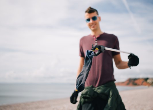 A man holding up a dark coloured sock up to the camera. He is using a litter picker to hold it. The sock is dark in colour and covered with sand.