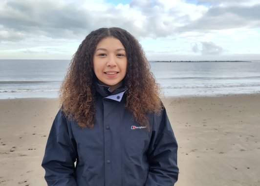 Trustee Rebecca smiles on a sandy beach with the ocean behind her. It looks like an overcast UK day. She has curly brown hair and is wearing a navy waterproof jacket.