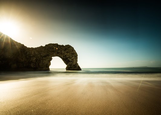 A long exposure photograph of the natural limestone arch known as Durdle Door on the Jurassic Coast in Dorset, England, with the sun low in the sky and blurred waves on the sand.