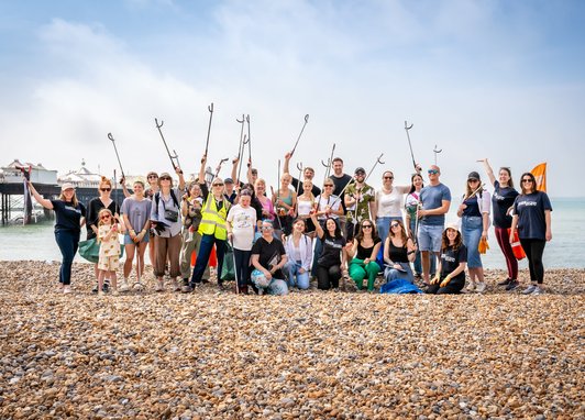 A large group of approximately 30 volunteers participating in a beach clean event stand smiling on a pebble beach, many holding litter pickers and bags, with a pier visible in the background.