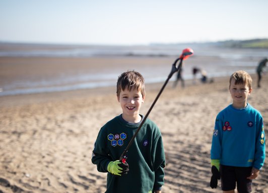 2 boys dressed in the Scouts uniform standing on a beach. One boy is holding up marine litter with a litter picker.