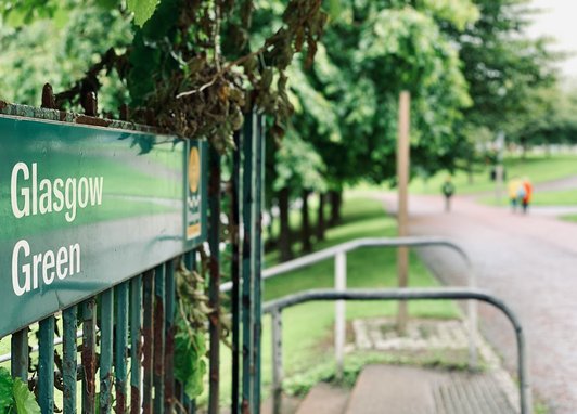 A close-up of a green metal sign that reads "Glasgow Green" in white lettering, attached to a weathered green iron fence. Behind the sign, a paved pathway leads into a lush, tree-lined park where blurred figures can be seen walking in the distance.
