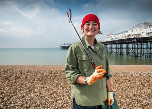 Zoe Lyons stands smiling on Brighton beach with the pier in the background. She is holding a litter grab over her shoulder and is wearing a khaki shirt and red beanie hat.