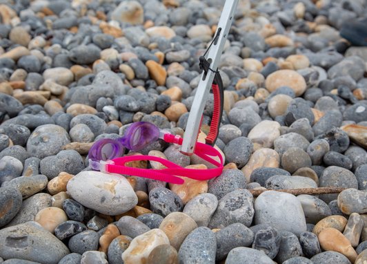 A pair of pink swimming goggles being held up by a litter picker on a pebble beach