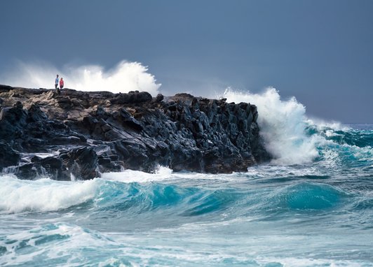 A shot of waves crashing into a cliff.On the cliff, 2 people can be seen.