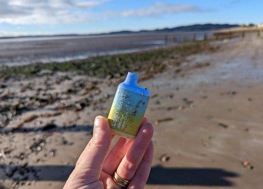 A hand is holding up a colourful disposable vape, the sandy beach is in the background. The disposable vape is litter.