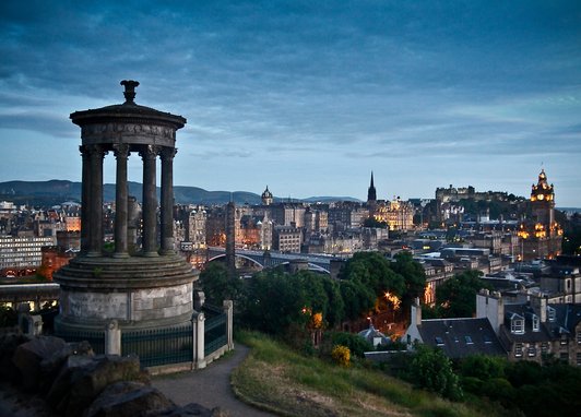 An evening view of the Edinburgh skyline from Calton Hill, featuring the Dugald Stewart Monument in the foreground and Edinburgh Castle illuminated in the distance under a twilight sky.