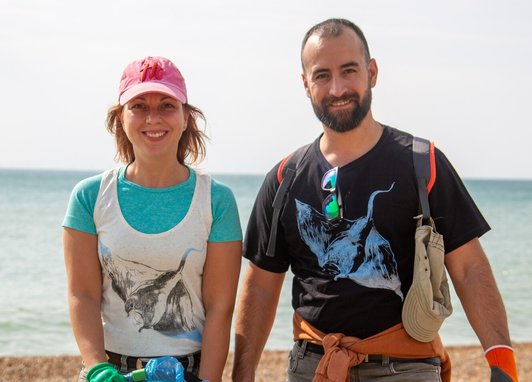 A man and a woman smiling at the camera on a shingle beach by the sea