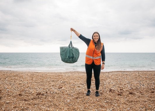 A woman in an orange hi-vis vest holding up a bag of litter with her right arm. She is standing on a pebble beach and is smiling.