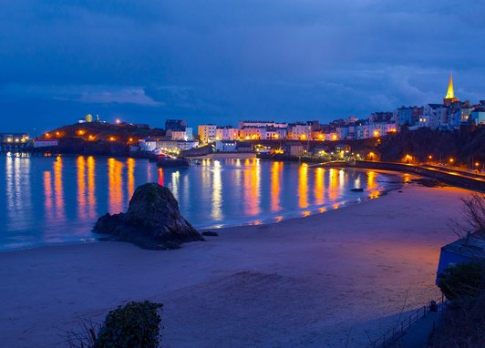 Tenby Harbour is captured at night time. The colourful lights from the buildings around the bay are reflected in the sea