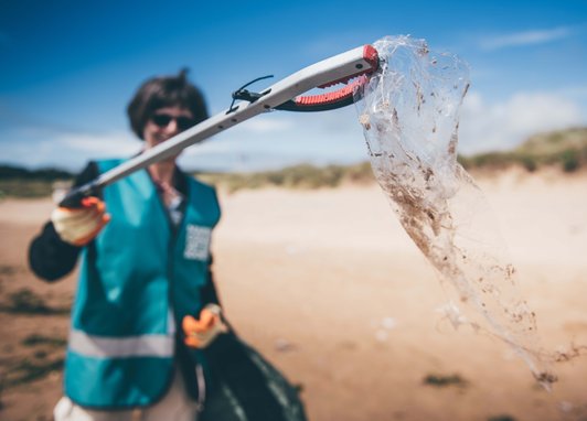 A woman holding up a piece of plastic wrapping using a litter picker to the camera. She is wearing a Marine Conservation Society vest. She is situated on a beach. The camera focus is on the wrapping making the woman blurry in the background.