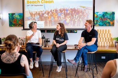 Zoe Lyons, a Marine Conservation Society staff member, and Cully from Cully and Sully, sit on stools in front of a large screen, giving a talk to an audience. All three people are smiling broadly.
