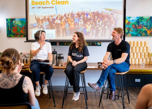 Zoe Lyons, a Marine Conservation Society staff member, and Cully from Cully and Sully, sit on stools in front of a large screen, giving a talk to an audience. All three people are smiling broadly.