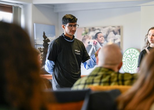 A young adult standing in front of some seated adults. He is wearing a Marine Conservation Society t-shirt and is speaking to the audience. Another young adult is standing near them listening to their speech.
