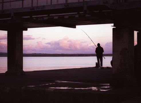 A silhouette of a lone fisherman under a pier.
