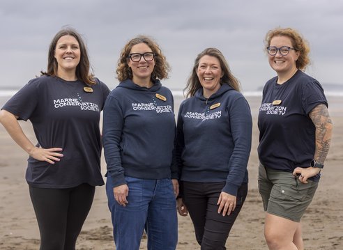 Four Marine Conservation Society staff stand together on a beach, smiling facing the camera, as they get ready to take part in a beach clean