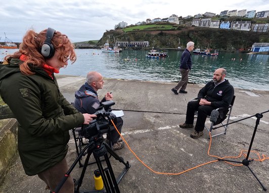 A film crew recording an interview with a man by the water in a busy harbor setting for the Valued Seas project
