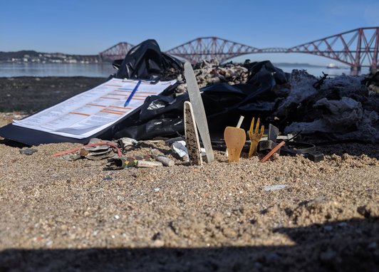 Single-use plastic on some sand. Next to it is a Marine Conservation Society beach clean survey form. The Forth bridge in Scotland can be seen behind it.