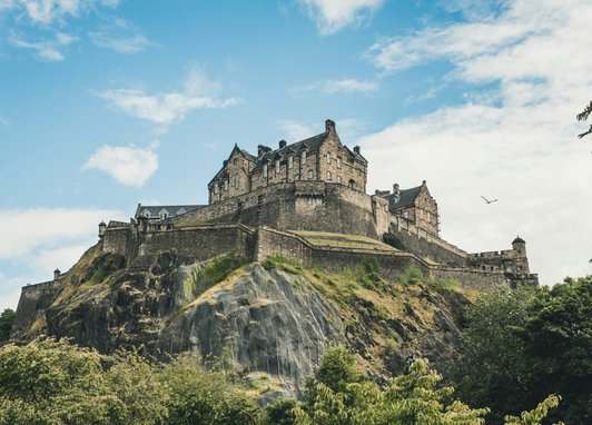 A low-angle shot of the historic Edinburgh Castle perched atop a jagged, rocky cliff in Scotland. The fortress stands prominently against a bright blue sky with wispy white clouds. Foliage frame the bottom of the image.