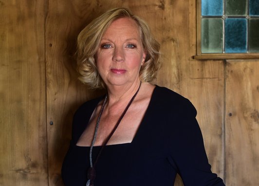Deborah Meaden smiles at the camera in what looks like the industrial backdrop of the Dragon's Den studio. She is wearing a smart black dress.