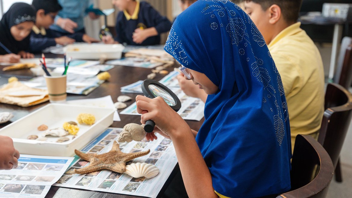 A girl in a blue hijab using a magnifying glass to examine a shell during an educational workshop on marine life.