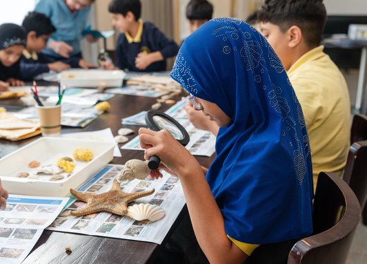 A girl in a blue hijab using a magnifying glass to examine a shell during an educational workshop on marine life.