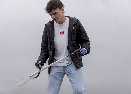 A teenage boy picking up marine litter with a litter picker. He is wearing a black coat and white t-shirt