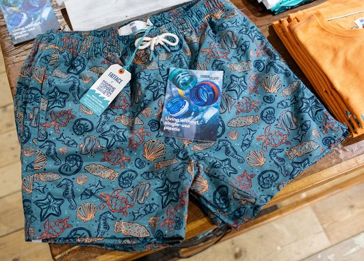 A pair of blubeach shorts patterned with outlines of turtles, seahorses, shells, starfish and types of fish. The label reads "Fatface X Marine Conservation Society". On top of the shorts is a leaflet that reads "Living without single-use plastic"