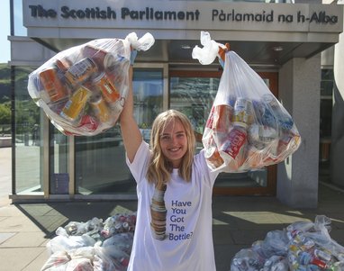 A female stands facing the camera wearing a white t-shirt that says "Have you got the bottle?". She has her arms raised and she is holding two bags filled with plastic bottles