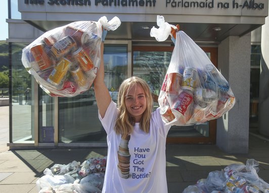 A female stands facing the camera wearing a white t-shirt that says "Have you got the bottle?". She has her arms raised and she is holding two bags filled with plastic bottles