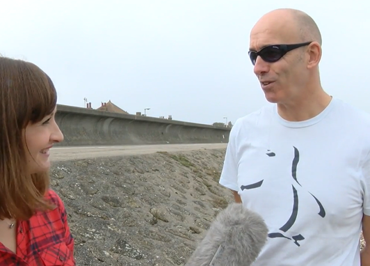 A man is being interviewed by a female presenter on a beach. They are near a concrete sea wall. The microphone can be seen at the bottom of the image.