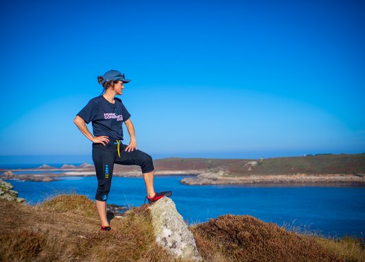 A woman with one leg resting upon a rock. She is overlooking a body of water with her hands on her hips. She is wearing a cap and a Marine Conservation Society t-shirt.